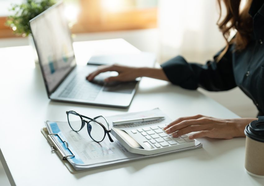 A property manager works on a calculator and laptop at a desk, with their glasses sitting folded in front of them.